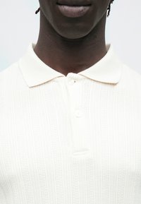 Polo creme em malha com padrão texturizado, gola clássica e dois botões no decote. Tecido suave com brilho subtil.