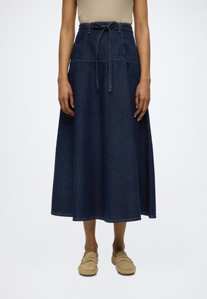 Hoch taillierter dunkelblauer Denim-Midirock mit gebundenem Gürtel, getragen mit beigefarbenen Loafers und einem neutralen Oberteil, gezeigt vom Oberkörper bis zu den Füßen.