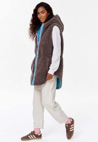 Fleecová vesta s kapucí, šedý povrch, jasně modrá podšívka, přední kapsy a plné zipové zapínání. V kombinaci se smetanovými kalhotami a hnědými teniskami.