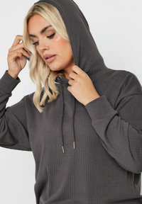 Grå ribbad hoodie med dragsko på huvan och långa ärmar, med en avslappnad passform och texturerad tyg, visad på en modell.