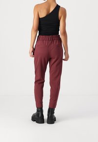 Burgundy tapered byxor med en texturerad yta, elastisk midja och två bakfickor, kombinerat med en svart topp med en axel och svarta klumpiga stövlar.