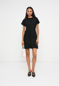Vestido negro de manga corta con textura y corte recto, que presenta una costura central. Combinado con zapatos negros de punta y un pequeño bolso rosa.