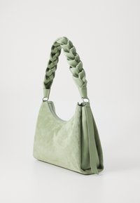 Saco de pele verde com uma alça trançada, apresentando um acabamento texturizado e suave. Fecho com zip e realces contrastantes lisos. Exibido em vista lateral.