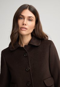 Cappotto di lana marrone con colletto arrotondato, cinque bottoni scuri e due tasche laterali. Tessuto testurizzato con una finitura liscia.