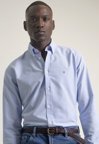 Camisa azul clara de botones, hecha de algodón, con mangas largas, tapeta delantera y un pequeño logo en el lado izquierdo del pecho, combinada con un cinturón trenzado marrón.