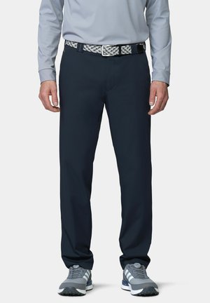 Man wearing navy blue pants, gray dress shirt, patterned belt, and gray athletic shoes standing with hands relaxed by sides.