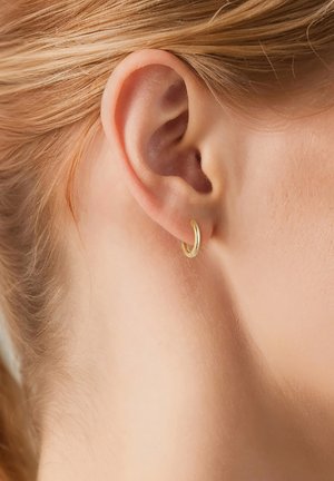 Boucle d'oreille en or attachée à l'oreille, présentant une surface lisse et polie avec une forme circulaire. Le teint de la peau est visible autour de la boucle.