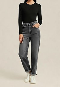 Femme portant un haut noir à manches longues, un jean gris taille haute avec revers, et des baskets blanches, debout devant un fond uni.