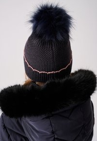 Neuletuttu tummansininen pipo, jossa on suuri tekoturkistupsu ja vaaleanpunainen korostevyö. Päällä musta turkisvuorinen takki. Karkea, lämmin materiaali.