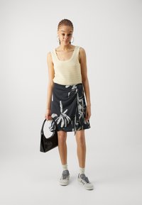 Strikket tanktop i kremfarge med bølgede kanter, parret med et svart omslagskjørt med palmebladmønster, mens hun holder en liten svart håndveske.