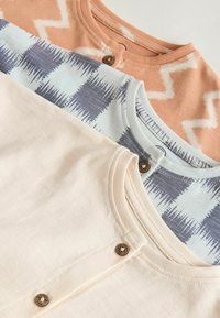 Trije zloženi otroški bodiji: breskvaste barve z vzorcem cik-cak, modro-sivo karo ter enobarvni krem, vsak z okroglim izrezom in gumbnimi poudarki.