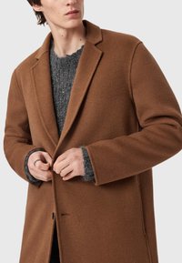 Smeđi vuneni kaput s izrezanim reverima, glatke teksture i jednostrukim kopčanjem na dugme. Ispod se nalazi sivi pleteni pulover.