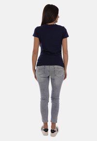 Donna con capelli lunghi e scuri che indossa una t-shirt blu navy, jeans skinny grigio chiaro e sneakers bianche, in piedi di spalle.