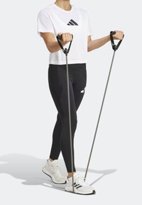 Leggings negras, camiseta blanca corta con logo negro y zapatillas deportivas blancas. Sosteniendo dos bandas de resistencia grises con mangos negros.