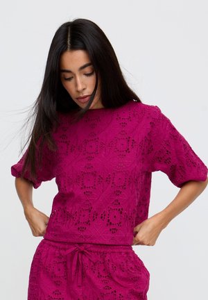 Junge Frau, die ein ärmelloses Oberteil und passende Shorts mit geometrischem Spitzenmuster in lebhaftem Magenta vor einfachem Hintergrund anpasst.