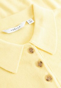 Polo en tricot jaune avec un col classique, doté de trois boutons marron et d'une étiquette "NOELLA" à l'intérieur du col.