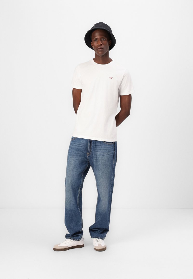 Weißes T-Shirt mit kurzen Ärmeln und kleinem rotem Logo, kombiniert mit locker sitzenden blauen Jeans und weißen Sneakers mit braunen Akzenten. Schwarze Bucket Hat.