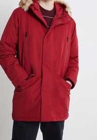 Röd parkasjacka med foder av konstpäls i huvan, dragkedjefickor och en slät tygfinish. Har en tryckknappsstängning framtill.