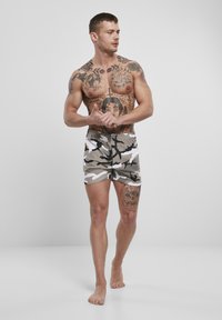 Bikini hlače v kamuflažni vzorec v bež, beli in črni barvi. Elastični pas z vrvico. Model stoji bos, pri čemer prikazuje tetovaže na rokah in trupu.