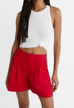 Młoda osoba z długimi warkoczami, ubrana w białą, bezrękawnikową, wiązaną na supeł bluzkę crop top i czerwone, plisowane szorty z wysokim stanem na białym tle.