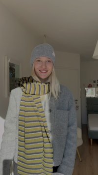 Joven sonriente en el interior con un gorro de punto gris, una chaqueta gris y una bufanda a rayas amarillas y grises alrededor del cuello.
