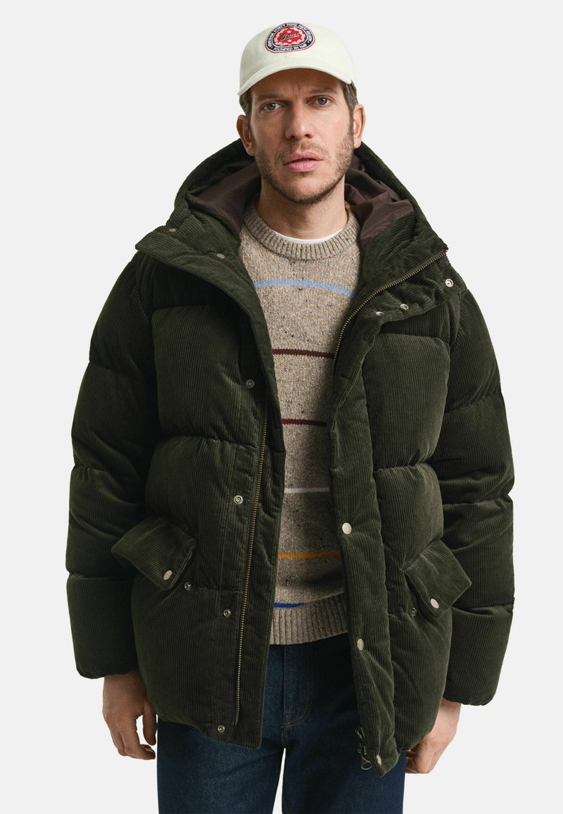 Grüne Cord-Pufferjacke mit Kapuze, ausgestattet mit großen Taschen und Druckknopfverschlüssen. Darunter ein brauner Pullover mit bunten Streifen.
