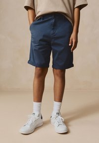 Pantalones cortos azul marino con puños enrollados y bolsillos laterales, combinados con zapatillas blancas y calcetines hasta el tobillo, usados con una camisa beige de manga corta.