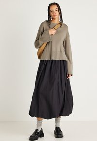 Jasnooliwkowy sweter z dzianiny o szerokich rękawach, w połączeniu z czarną, fakturowaną, spódnicą midi. Modelka nosi szare skarpetki i czarne buty na platformie.