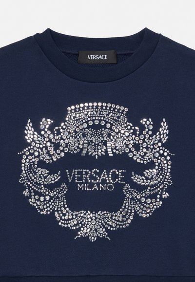 Tengerészkék ruhadarab ezüst strasszkő címer dizájnnal és a „VERSACE MILANO” szöveggel, amely egy lekerekített nyakkivágás alatt középen helyezkedik el.