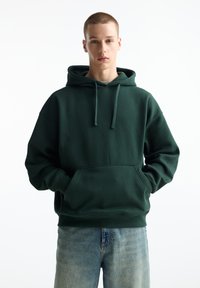 Dunkelgrüner Kapuzenpullover aus weichem Stoff, mit einer Vorderentasche, Kordelzugkapuze und gerippten Bündchen. Trägt eine helle blaue Jeans.