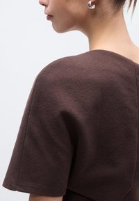 Καφέ κοντομάνικο τοπ με υφή. Στρογγυλή λαιμόκοψη και λεία ραφή στους ώμους. Ορατό ασημένιο σκουλαρίκι σε σχήμα κρίκου.