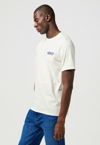 Camiseta de algodón crema con mangas cortas, con la palabra "HOWDY" en texto rojo y azul en el lado izquierdo del pecho. Combinada con jeans de mezclilla azules.