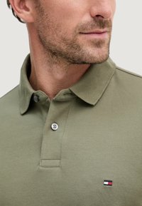 Polo verde oliva in tessuto strutturato, con colletto classico, due bottoni e un piccolo dettaglio brandizzato in rosso, bianco e blu.