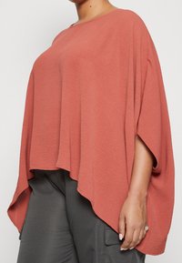 Terrakottafarbener Poncho mit lockerem Sitz, aus strukturiertem Stoff, mit weiten Ärmeln und asymmetrischem Saum. Kombiniert mit dunklen Hosen.