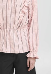 Rožinė, tekstūruota bluzė su vertikaliomis juodomis juostelėmis, raukšlėtais akcentais šoninėse dalyse ir surinktomis apykaklėmis ilgose rankovėse. Dėvima su juodais kelnais.