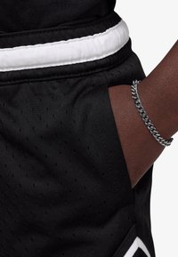 Black athletic shorts with a white elastic waistband. Features side pockets and a textured fabric. A silver chain bracelet is visible on the arm.