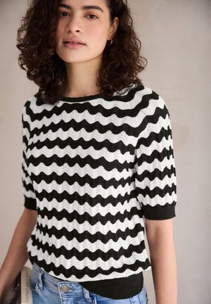 Žena s kudrnatými vlasy, která má na sobě černo-bílý svetřík s krátkými rukávy se vzorem cikcak a světle modré džíny, stojící před neutrálním pozadím.