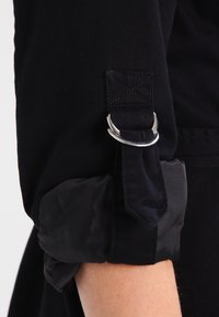 Czarna tkaninowa rękawka podwinięta i zabezpieczona czarnym paskiem oraz srebrną klamrą D-ring powyżej przedramienia.