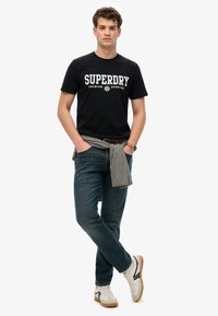 Sorte t-shirt med hvid "SUPERDRY" tekst, parret med blå jeans og en grå sweater bundet om livet. Læderur og hvide sneakers.