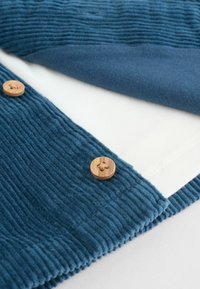 La tela de tergal azul presenta estrías verticales, complementadas por una suave superposición azul y dos botones de madera con acabado natural.