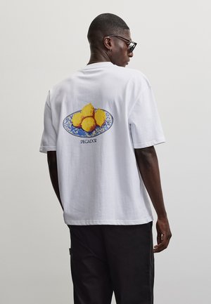 Mann trägt weißes T-Shirt mit blauem Teller, hält fünf gelbe Zitronen und hat den Schriftzug "PEGADOR" auf dem Rücken, schwarze Hose und Sonnenbrille.