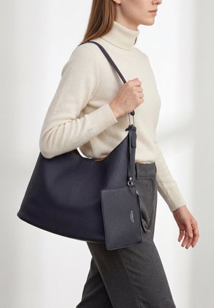 Frau in cremefarbenem Rollkragenpullover und grauer Hose trägt eine große dunkelblaue Leder-Umhängetasche mit passender angenähter Tasche.