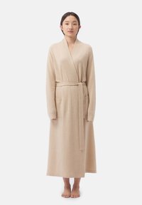 Robe longue beige en tissu doux ; dotée d'un col châle, de manches longues, d'une ceinture à nouer et de deux poches avant. Texture lisse, coupe jusqu'au sol.