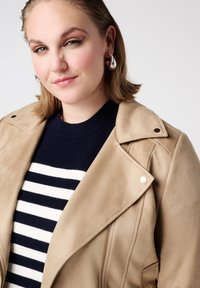 Tan suède jas met diagonale ritssluiting aan de voorkant, zilverkleurige accenten en een reverskraag, gedragen over een marineblauw gestreepte trui. Gladde textuur en een getailleerde pasvorm.