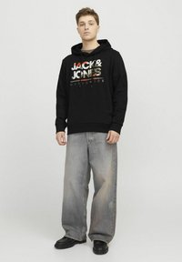 Melna kapuce ar grafisku druku gaišās krāsās un tekstu "JACK & JONES WORLDWIDE." Brīvas formas gaiši pelēkas džinsi un melnas kurpes.