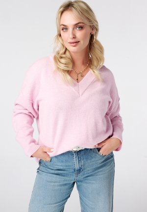 Vrouw met blond haar die een lichtroze V-hals trui draagt, blauwe spijkerbroek, gouden oorbellen in hoopvorm en meerdere kettingen, staand met handen in de zakken.