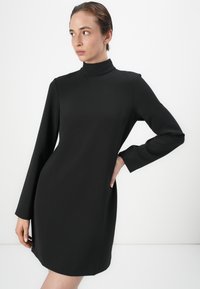 Robe noire à manches longues avec un col haut ; forme ajustée en tissu lisse et texturé ; longueur genou avec des détails de couture subtils.