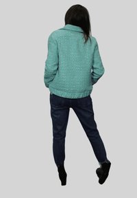 Chaqueta acolchada verde azulado con cuello, tela texturizada y bajo acanalado, combinada con vaqueros azul oscuro y botines negros.