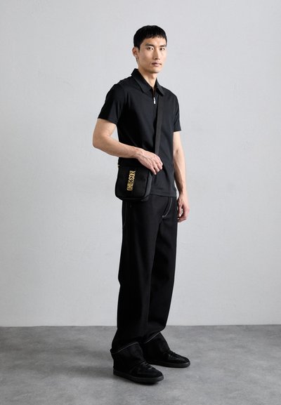 Polo negru cu guler cu fermoar, asortat cu pantaloni negri largi. O geantă de umăr neagră cu litere galbene se află pe umăr. Adidași negri.