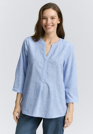 Chemise à manches longues bleu clair avec un décolleté en V, texture douce et motif à carreaux subtil. Coupe décontractée, portée par-dessus un pantalon en denim.
